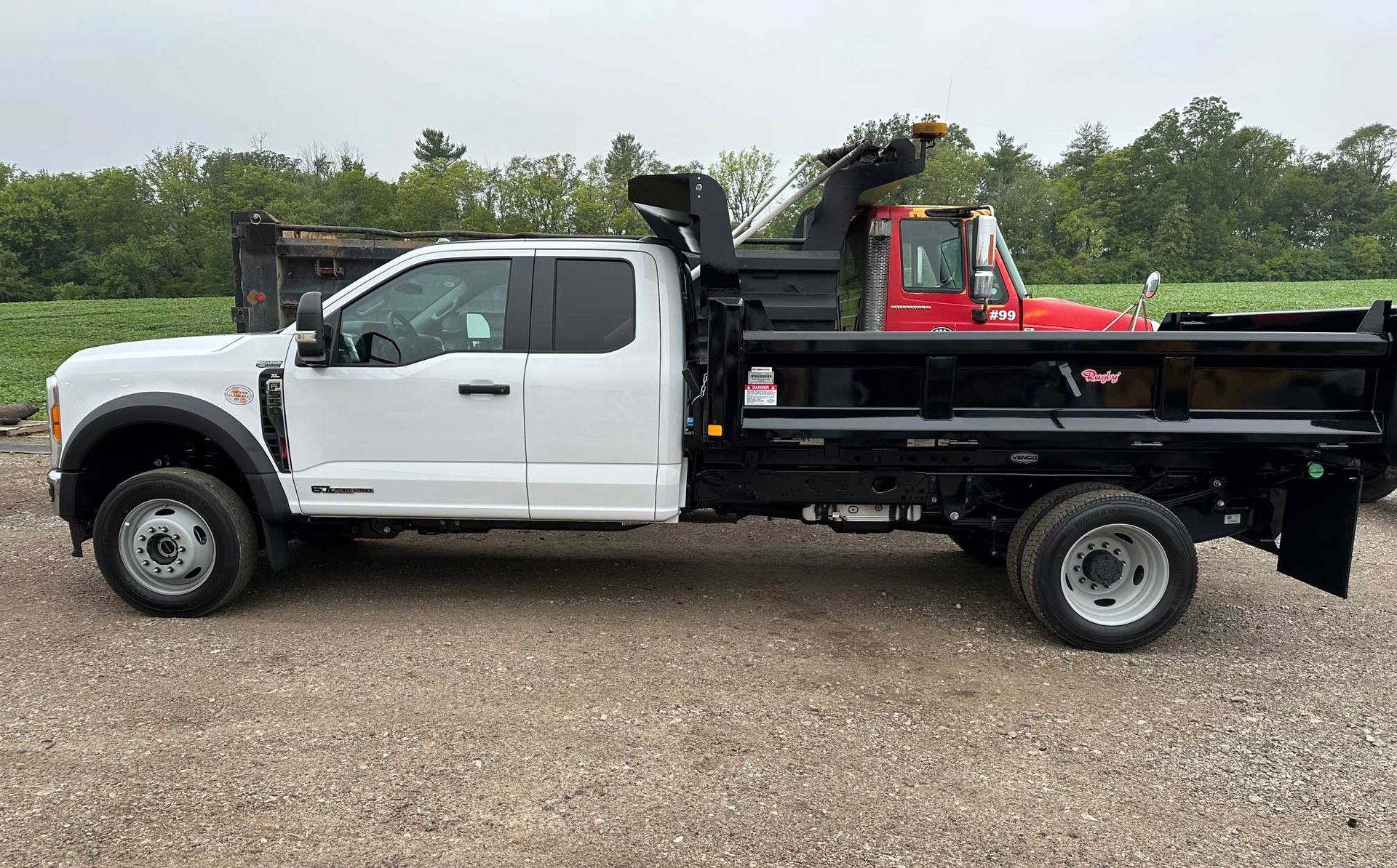White Ford F-550 dump truck with a black bed, parked on gravel. Red truck visible in the background.