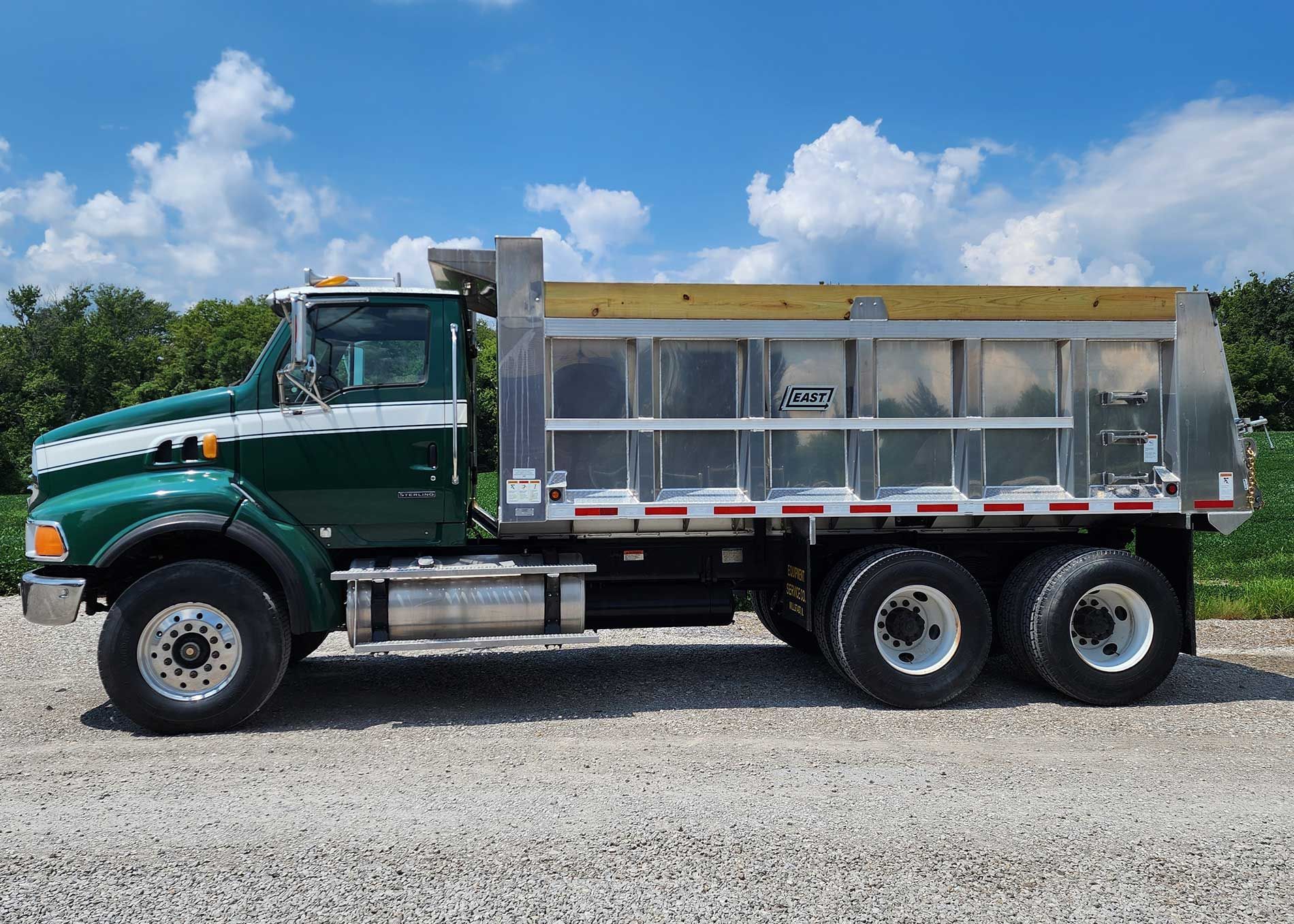 Green and white dump truck on a gravel road under a blue sky with clouds.