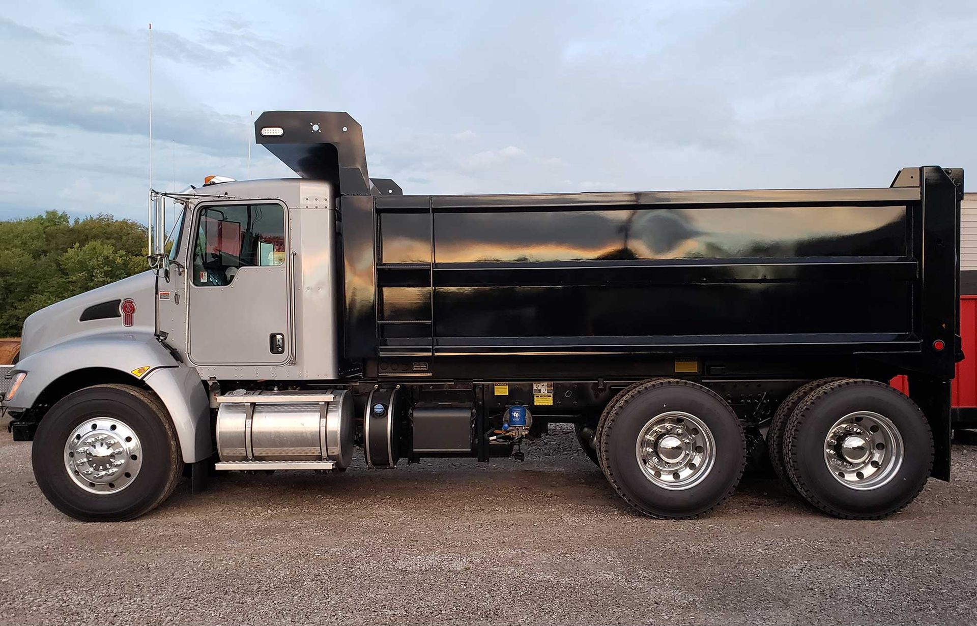 Silver and black dump truck on gravel, outdoors under a cloudy sky.