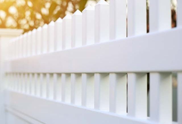 A close up of a white picket fence with trees in the background.