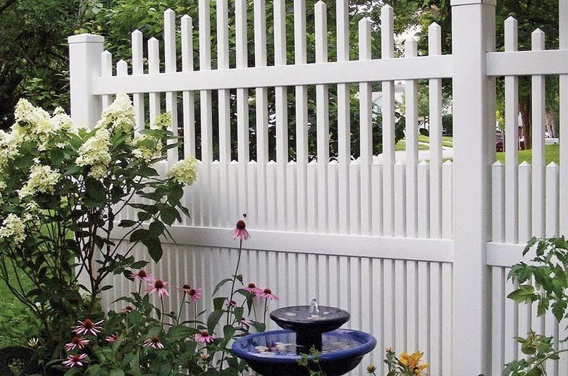A white picket fence surrounds a garden with flowers and a fountain.