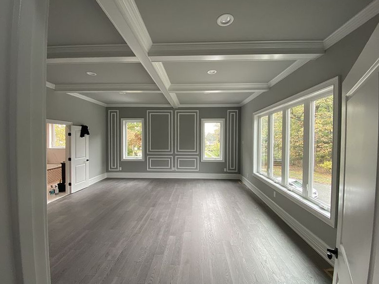 Empty room with gray walls, white trim, and wood flooring. Features include large windows and a coffered ceiling.