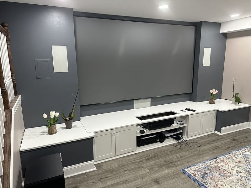 Home theater setup: gray screen, white cabinetry with entertainment equipment, gray walls, and light-colored flooring.