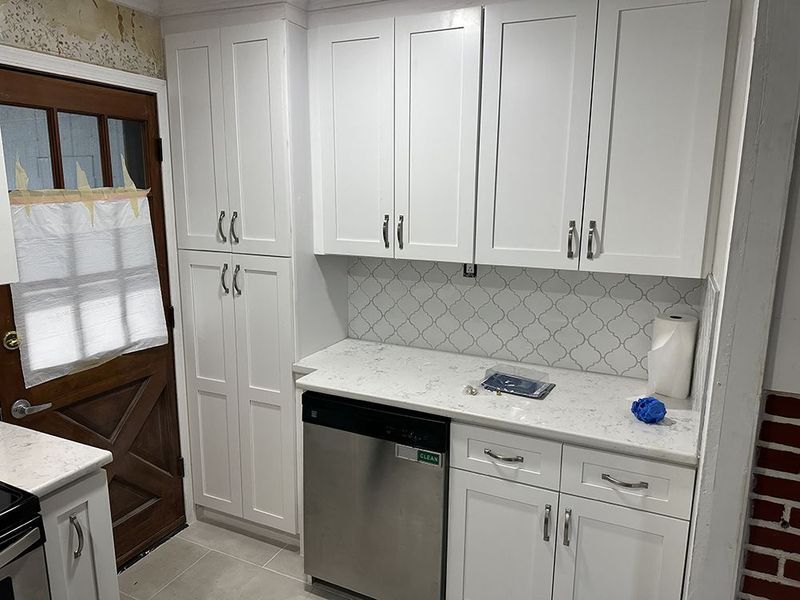 White kitchen cabinets with stainless steel dishwasher and white countertop; a wooden door is to the left.