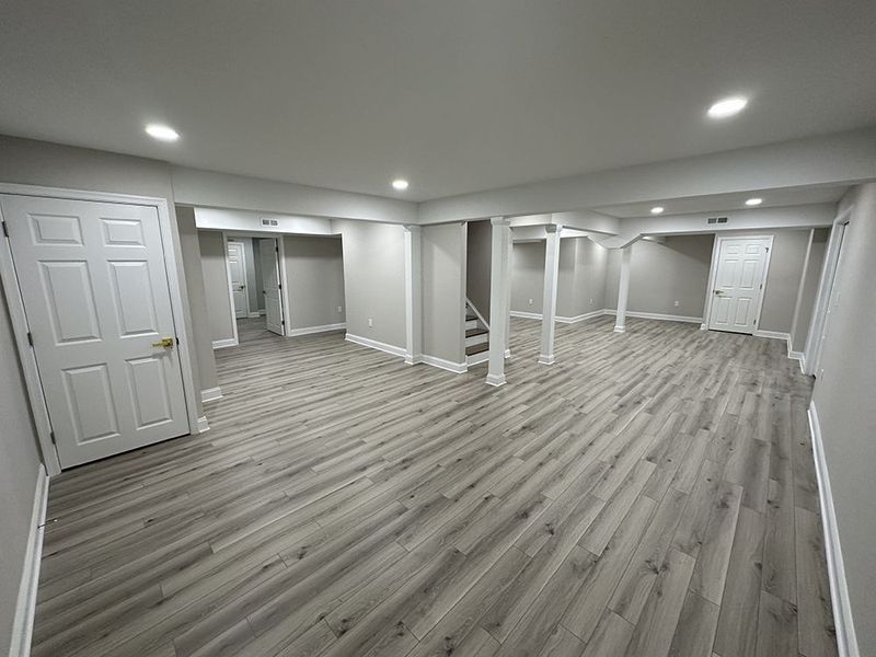 Finished basement with gray wood-look flooring, white doors, support columns, and recessed lighting.