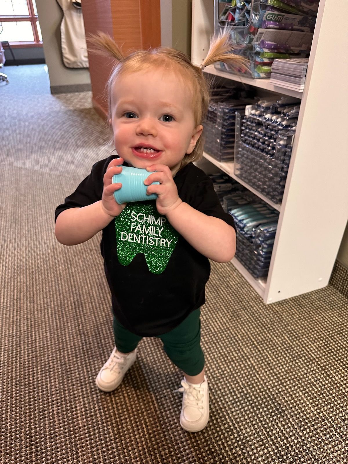 Smiling toddler with pigtails holding blue object, wearing a black shirt, green pants, and white shoes.