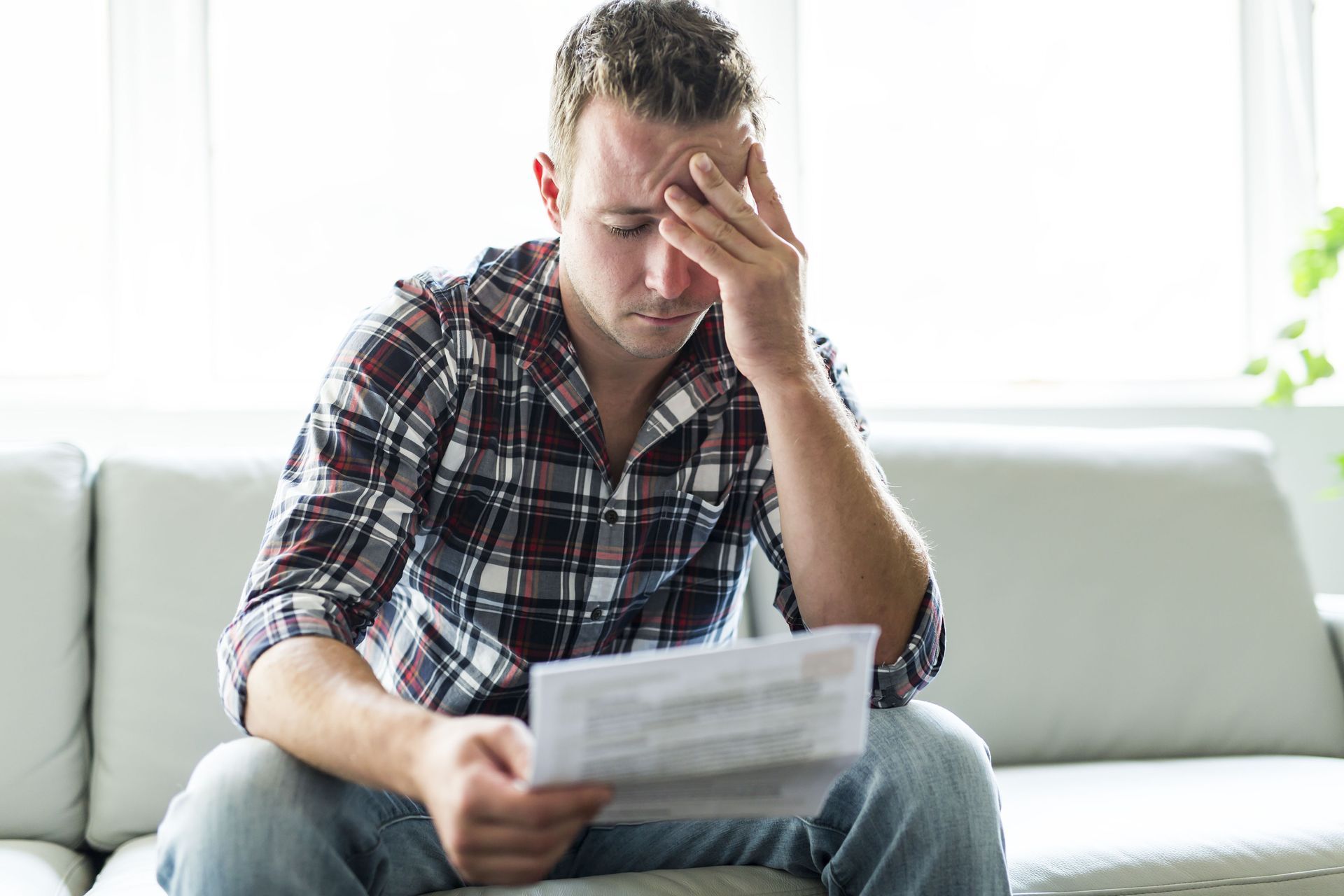 Man sits on a sofa, looking at papers with a stressed expression; he holds his head.