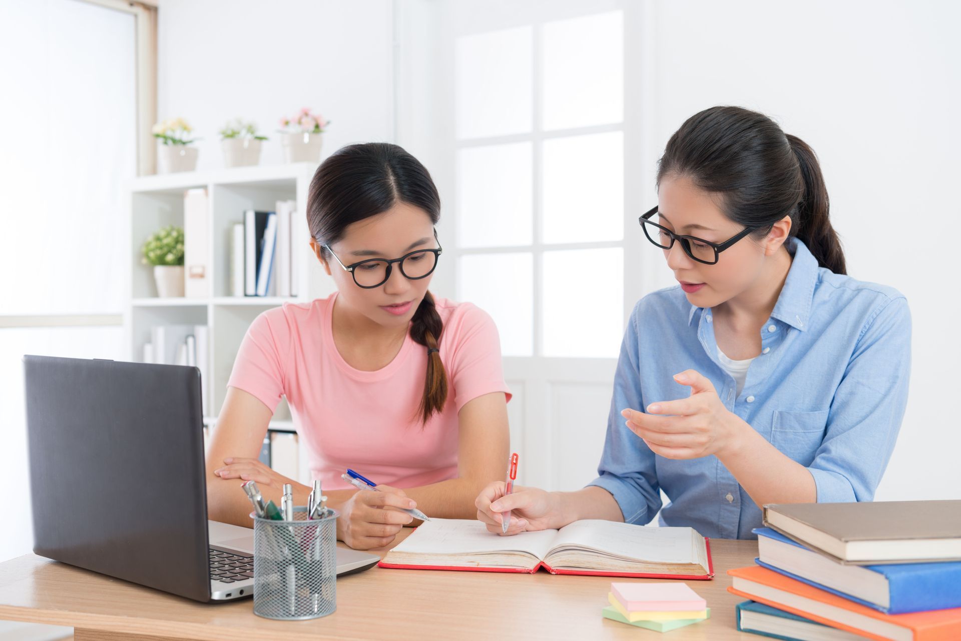 Two people wearing glasses at a desk studying, one pointing at notes while the other writes.