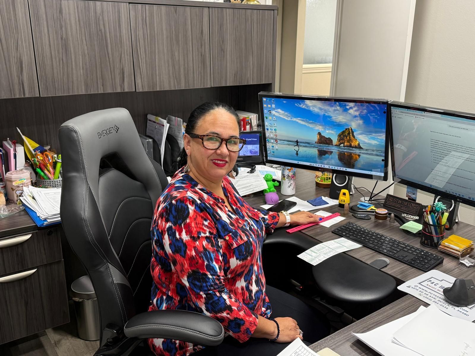 Woman at a desk with two monitors; wearing glasses and a patterned shirt, smiling. Office setting.