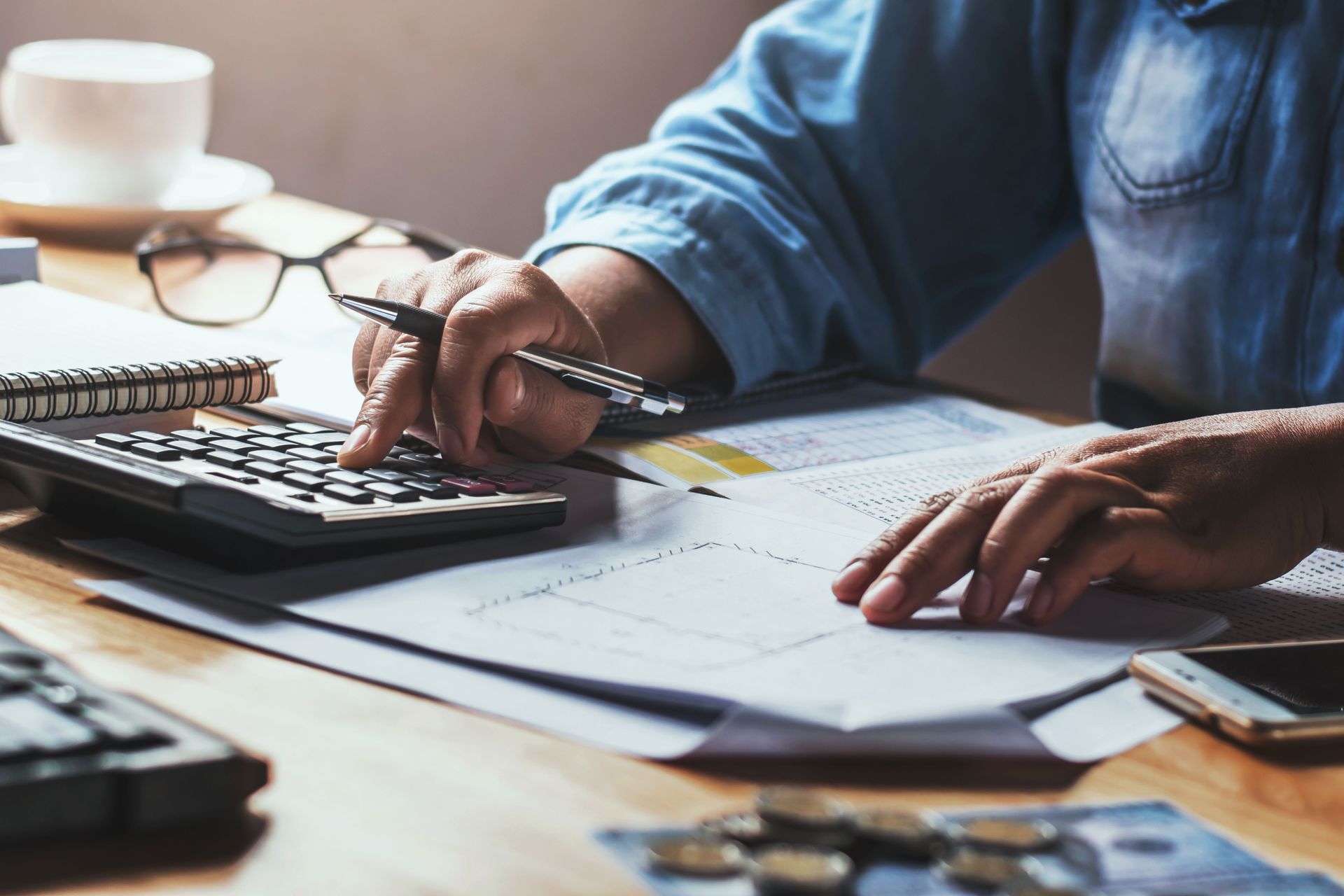 Hands using a calculator with paperwork, glasses, and coins on a desk.