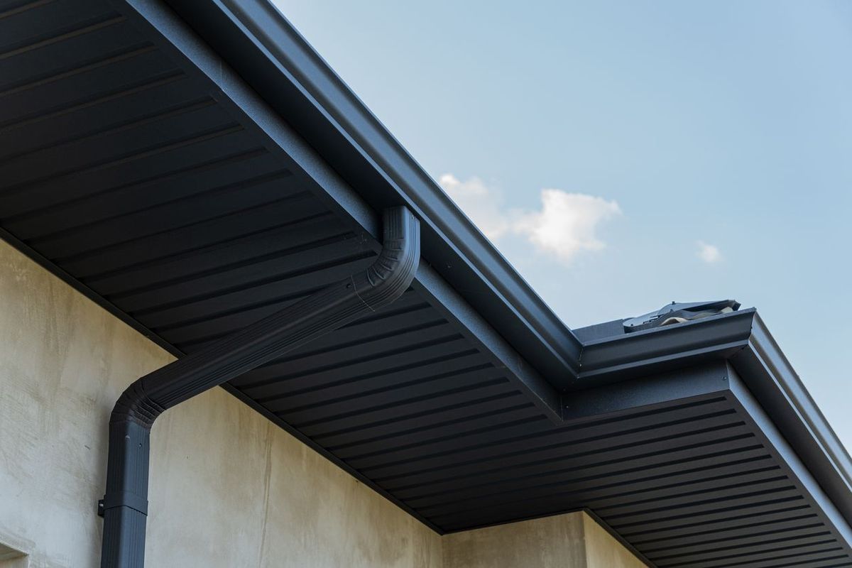 Dark gray rain gutter and downspout along the edge of a building against a blue sky.