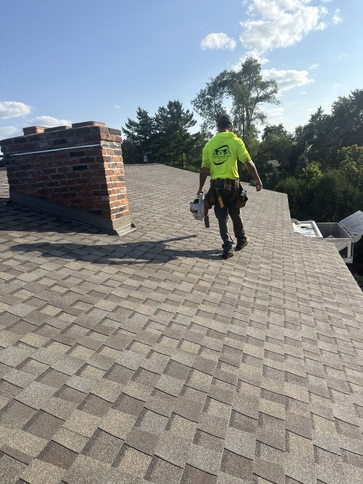 Roofer walking on a shingle roof, wearing a yellow shirt, with a brick chimney in the background.