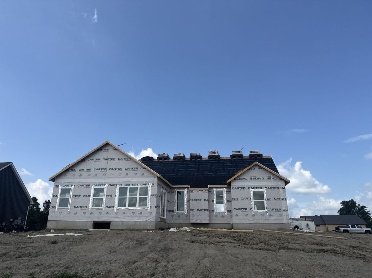 New house under construction against a blue sky; exterior walls wrapped; roof partly shingled.
