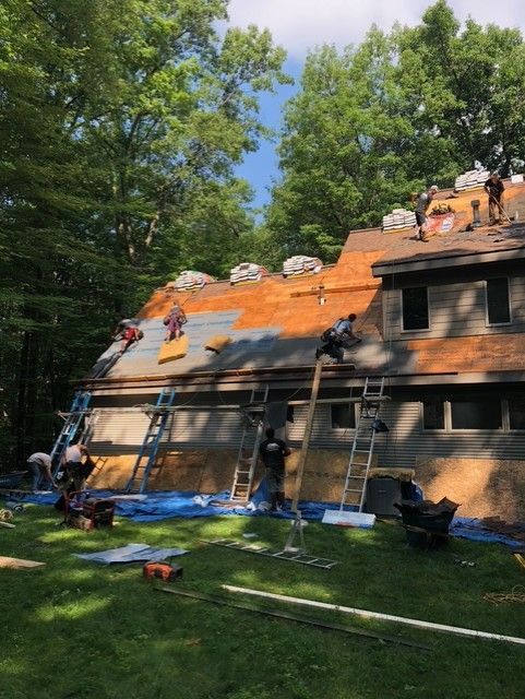 Roofing crew on a house, replacing shingles; ladders, tools, and tarp on the lawn, sunny day.