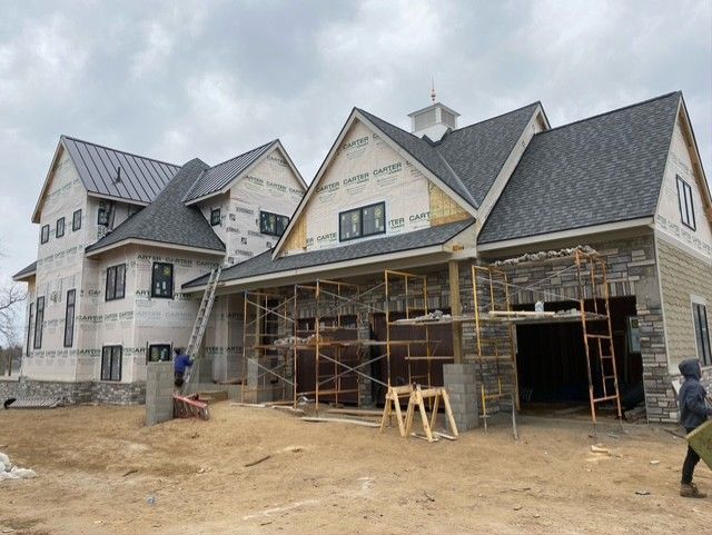 Construction site of a two-story house with scaffolding, stone facade, and dark gray roof on a cloudy day.