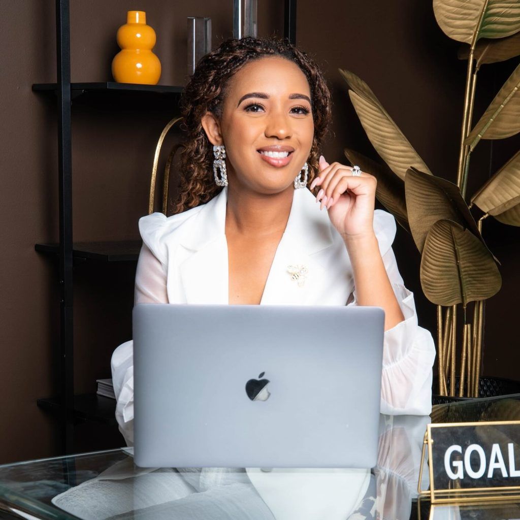 Woman in white blazer smiling, sitting at desk with laptop, "GOALS" sign.
