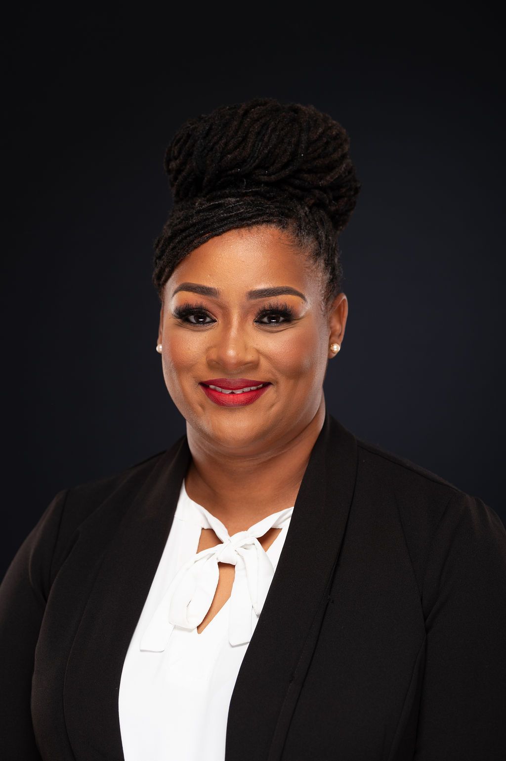 Woman in a black blazer and white blouse, smiling. Braided updo and red lipstick. Dark background.