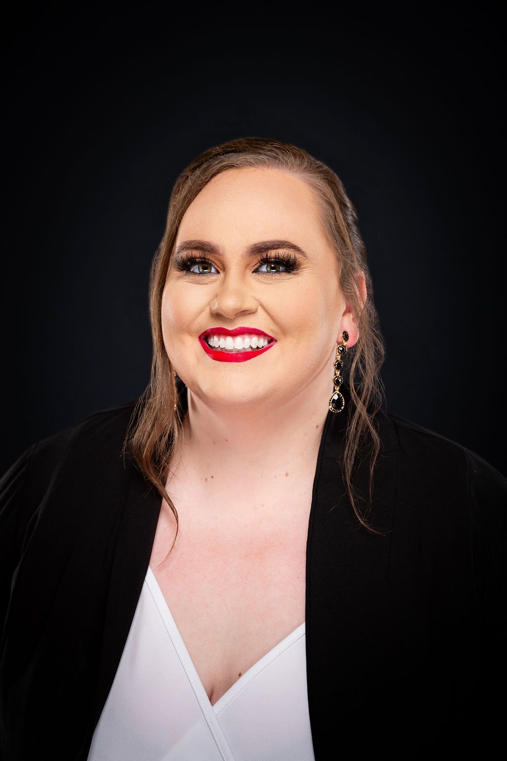 Woman with red lipstick smiles, wearing a black blazer over a white top against a dark background.