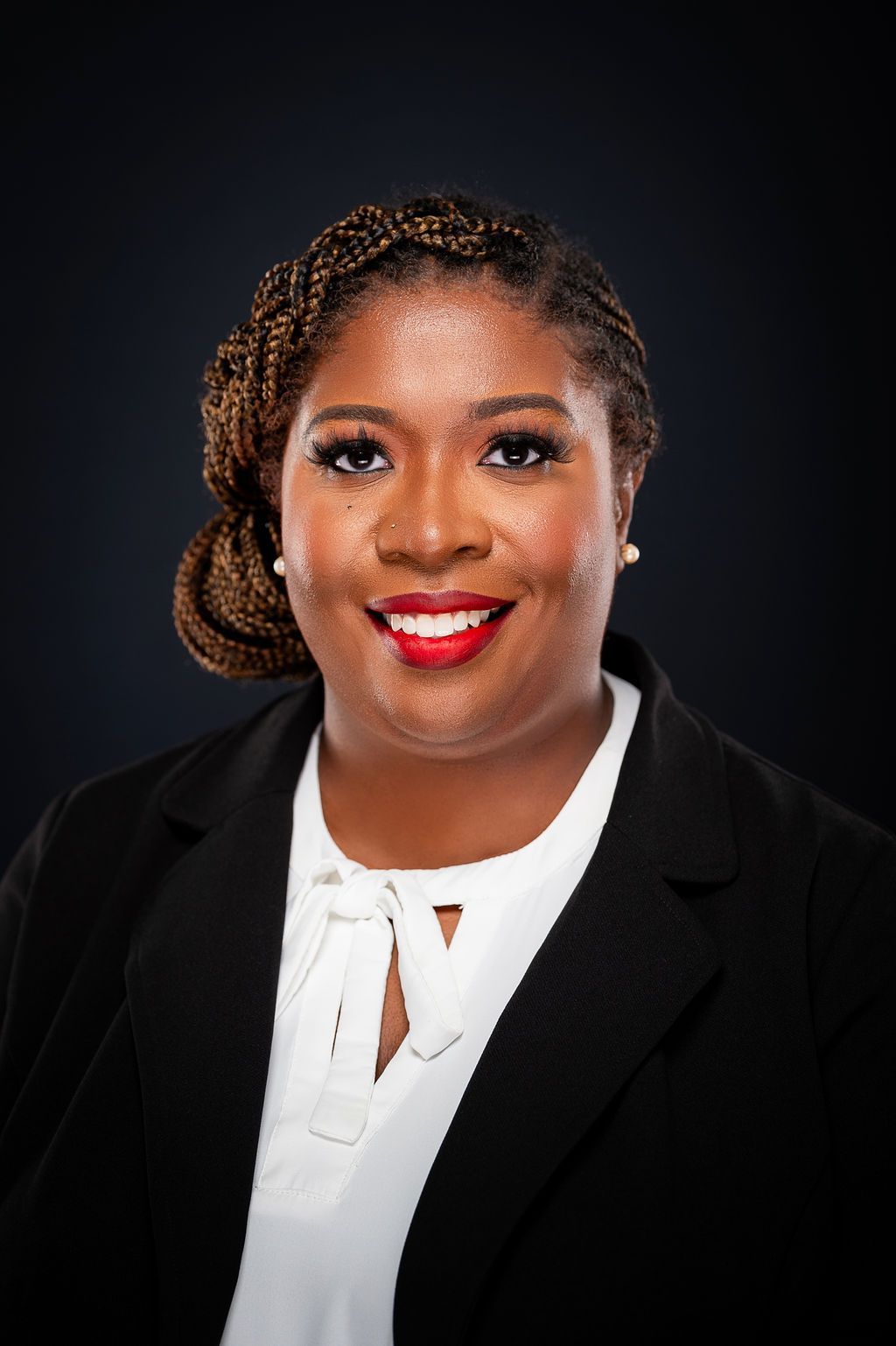 Woman in a black blazer and white blouse smiles, wearing braided hair, red lipstick, and pearl earrings.