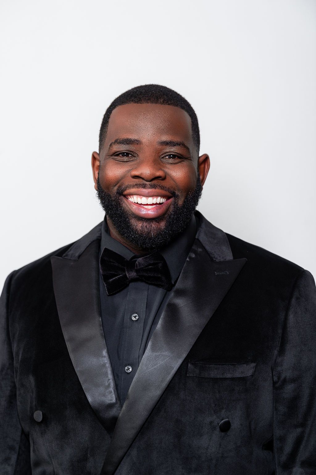 Man in black tuxedo smiles at the camera, against a white background.