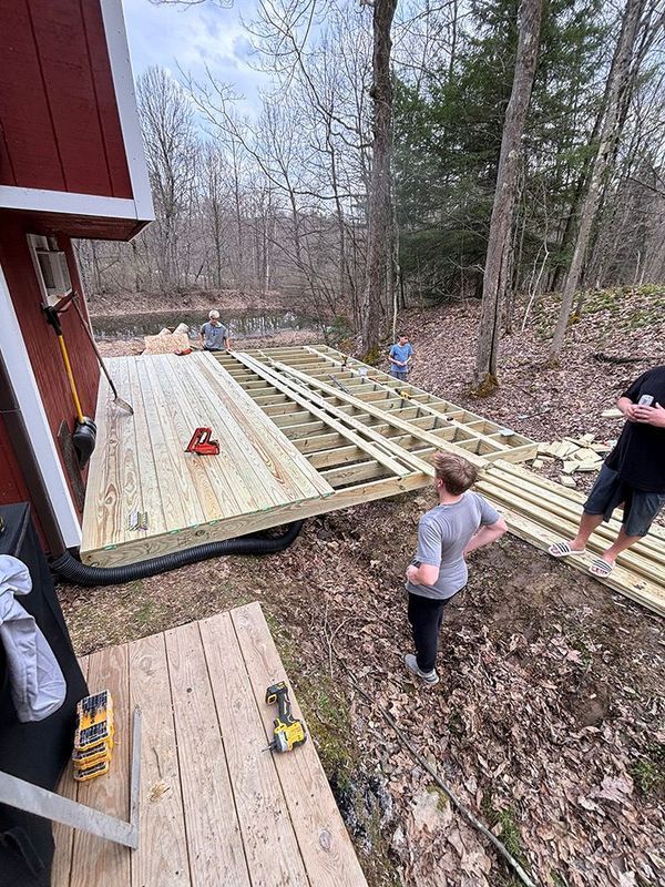 Construction of a wooden deck near a red building and pond, with several people working.