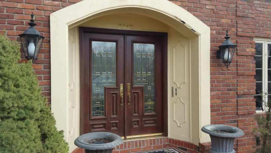Brown double doors with glass panels and decorative trim, under an arched entryway. Two black lanterns and planters flank the entrance.
