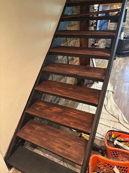 Metal staircase with dark wooden steps against a beige wall. Tools and a blanket are at the base.