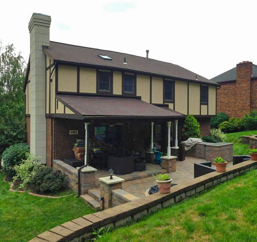 Two-story house with a brick patio and covered outdoor seating area. Brown and beige exterior. Green lawn.