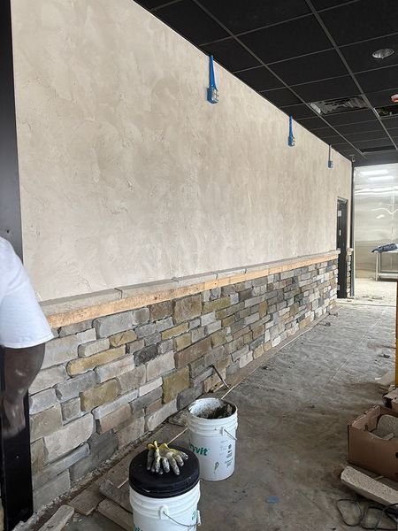 Interior wall with stone veneer installation; worker, buckets, exposed wiring, and unfinished ceiling.
