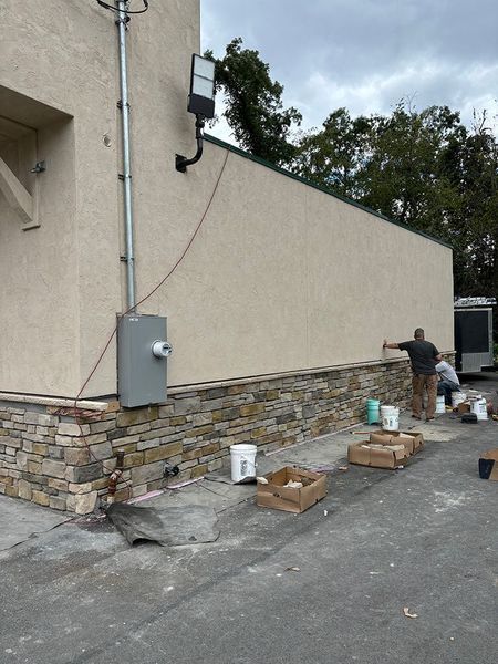 Exterior wall being worked on, with stone veneer at the base and stucco above. Two workers are applying the finishing touches.