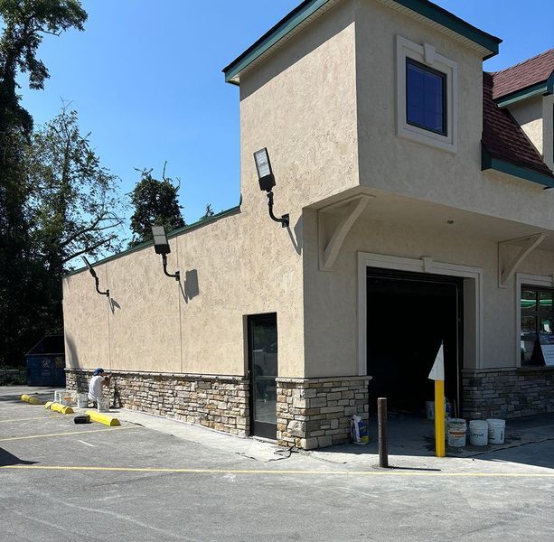 Building exterior with beige stucco, stone facade, black door, and parking lot. A person paints on the left.