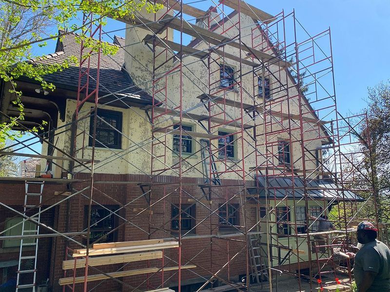 House under construction, scaffold surrounding exterior. Brick base, white stucco, and dark roof. Workers visible.