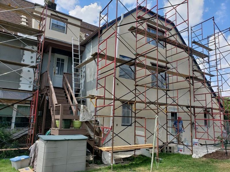 Building exterior under renovation with scaffolding. Workers, tan walls, red scaffolding, and green grass.