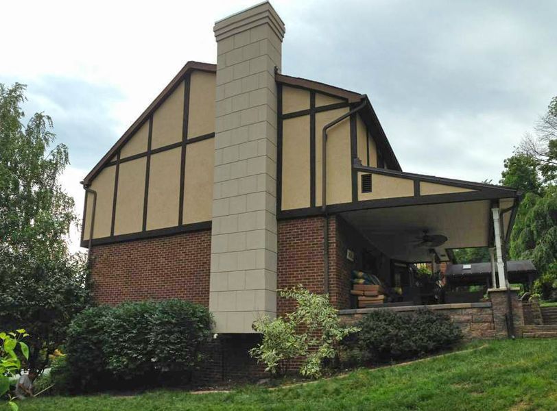 Tudor-style house with beige stucco and brown trim, brick base, and tan chimney. Covered porch and green landscaping.