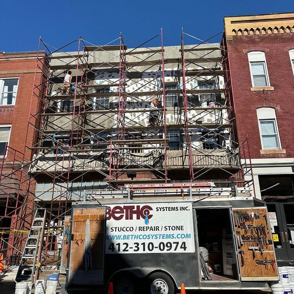 Building undergoing stucco repair with scaffolding; workers visible. Bethco Systems Inc. trailer with contact info in front.