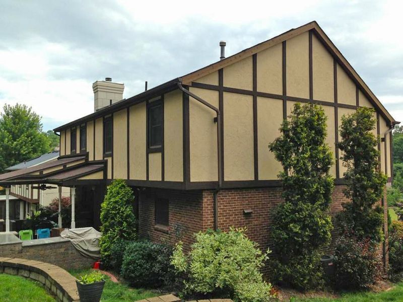 Two-story Tudor-style house with brown trim, tan siding, and brick base, surrounded by greenery.