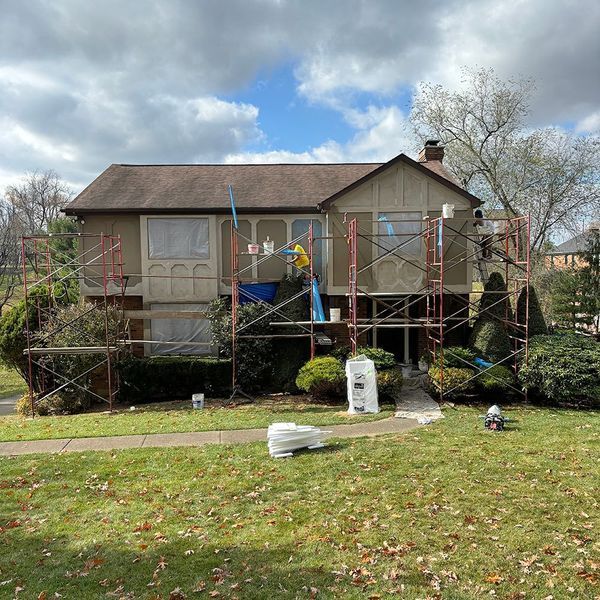 House exterior under renovation, with scaffolding and workers present. Green grass and cloudy sky.