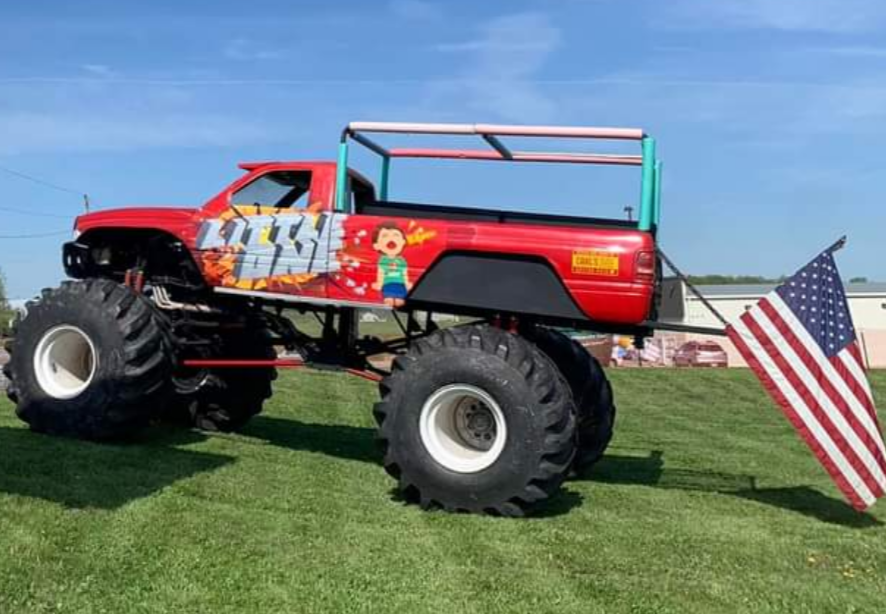 A red monster truck is parked in a grassy field next to an american flag.