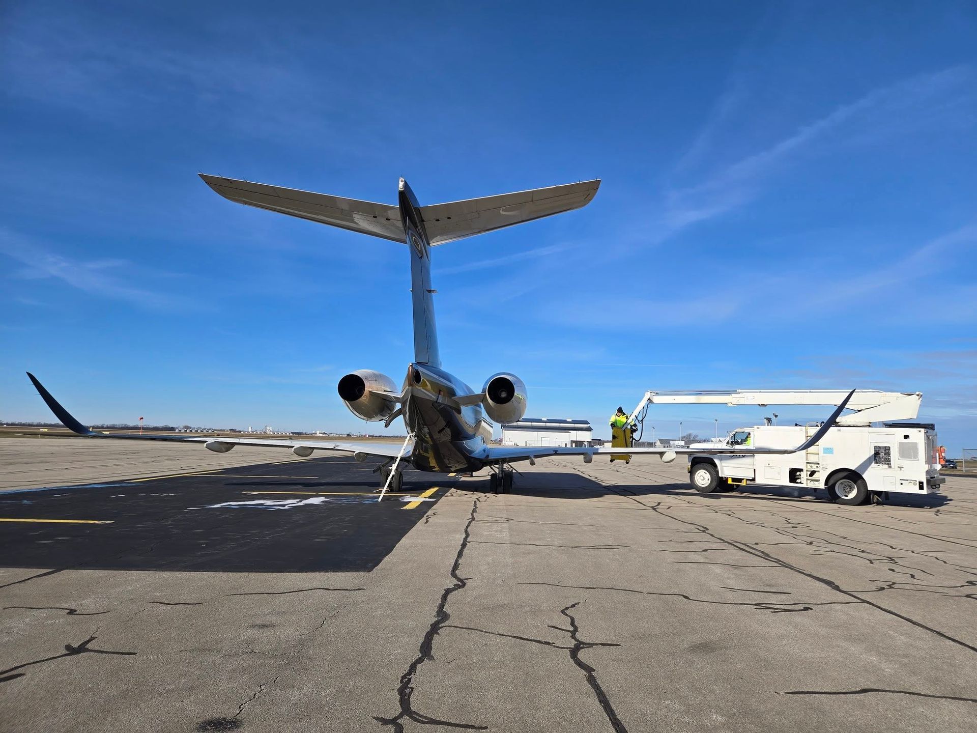 Jet on tarmac with tail raised, being serviced by a white truck with lift, under a clear, blue sky.