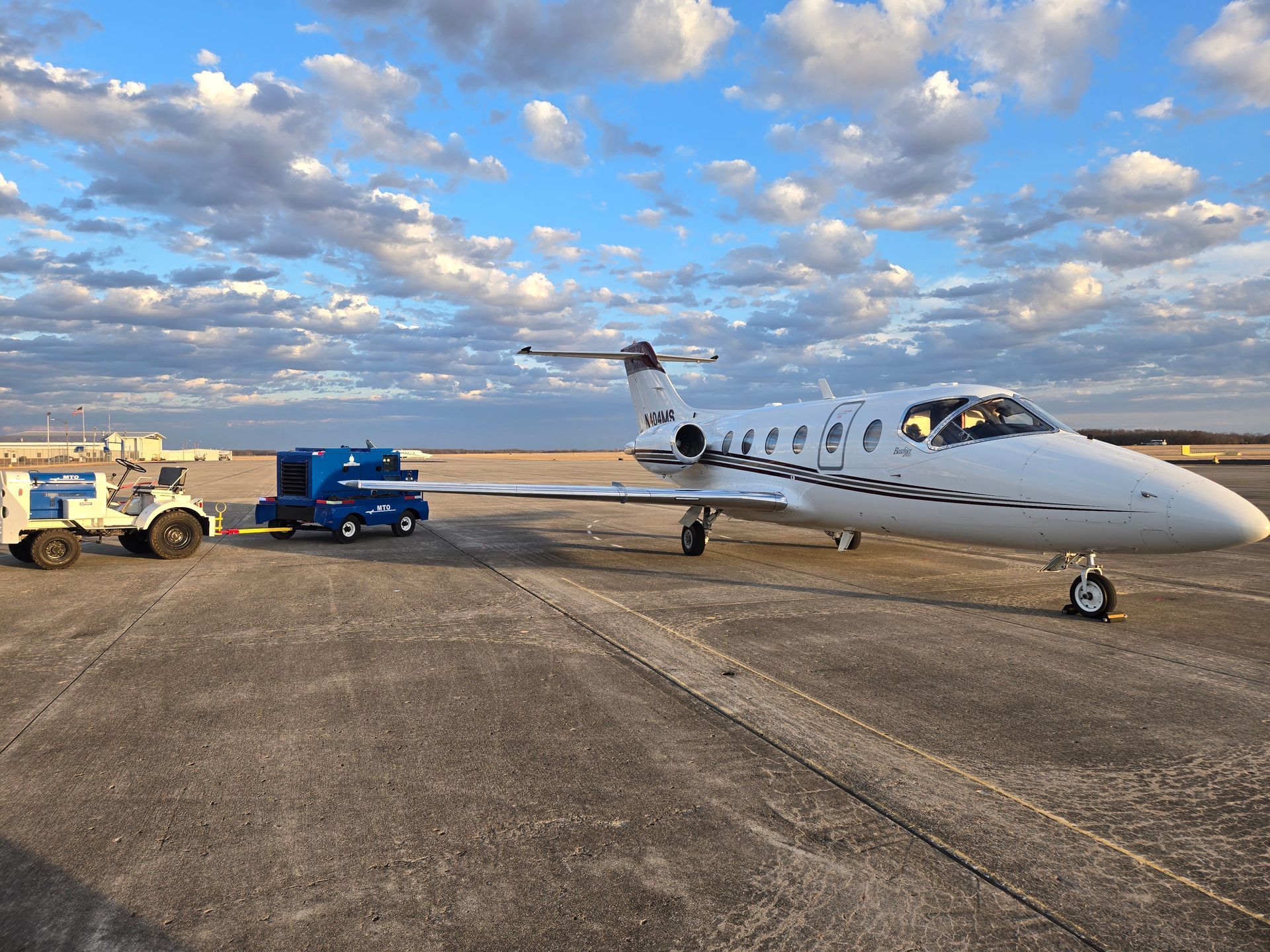 White private jet parked on tarmac with blue service vehicles under a cloudy sky.