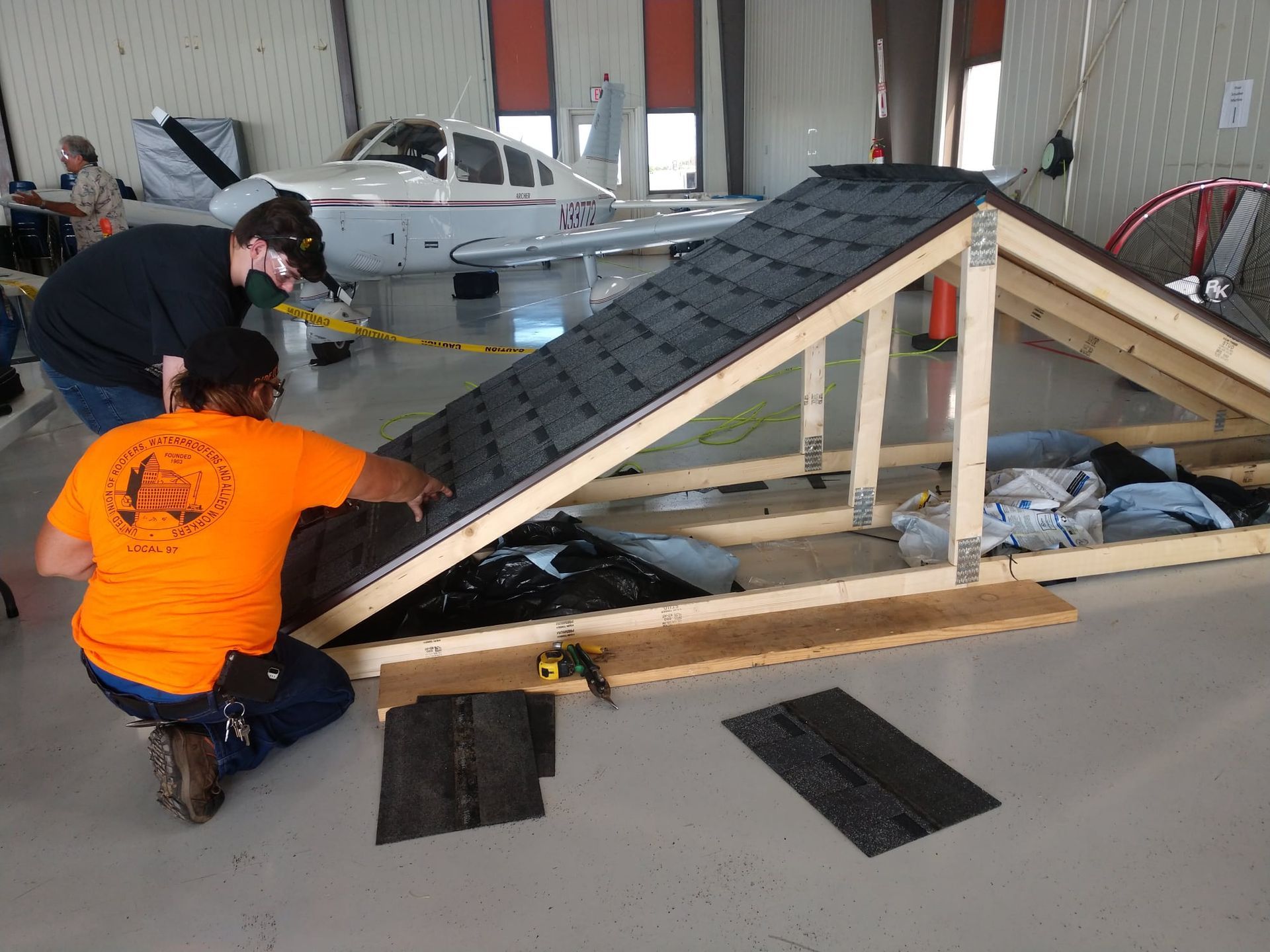Two men are working on a roof in a hangar with an airplane in the background.