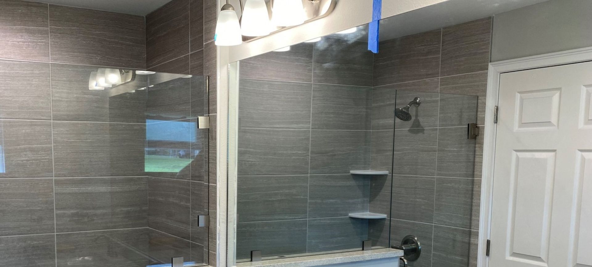Glass shower enclosure in a gray-tiled bathroom beside a white door.