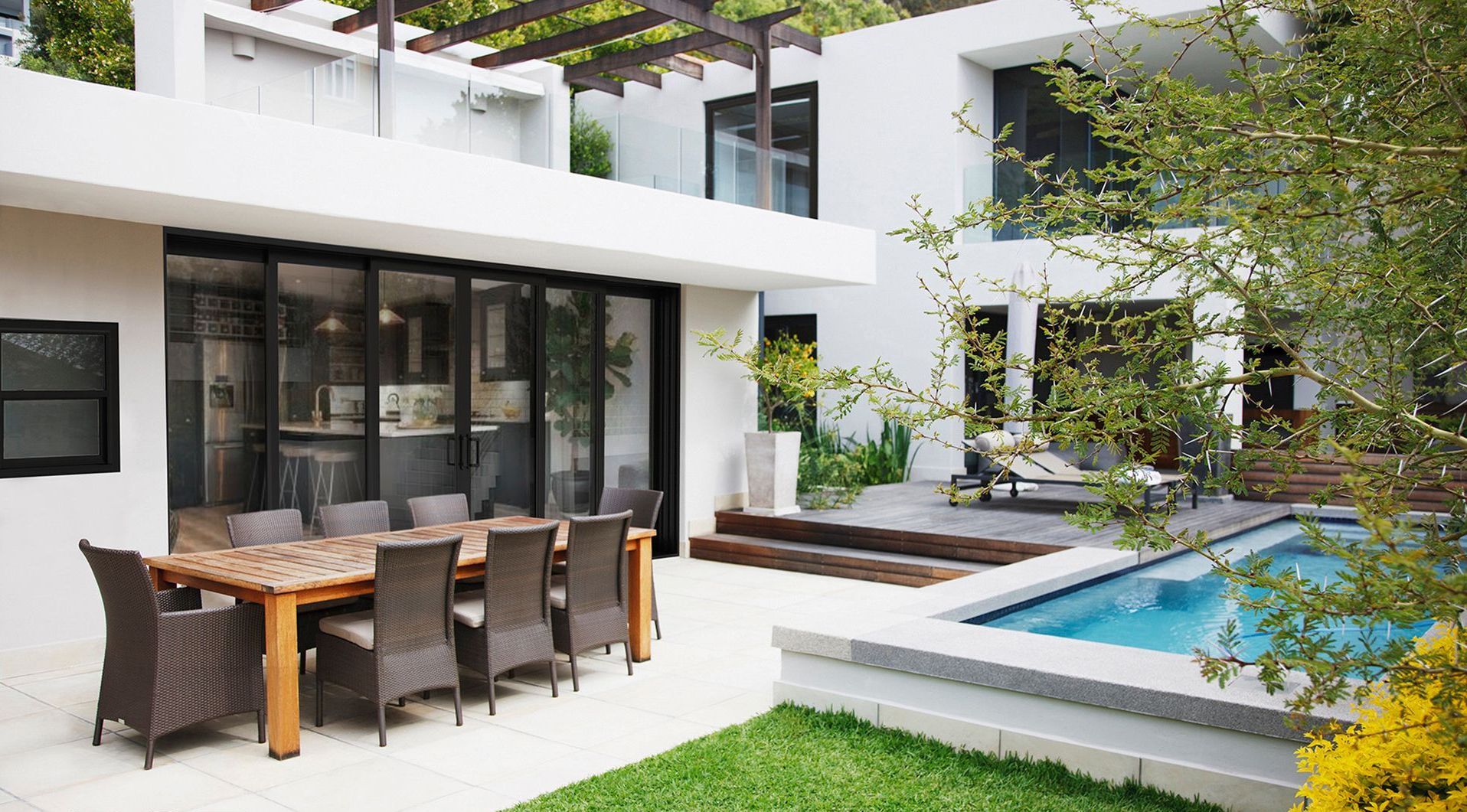 Modern patio with dining table beside a pool and white contemporary house with glass doors.