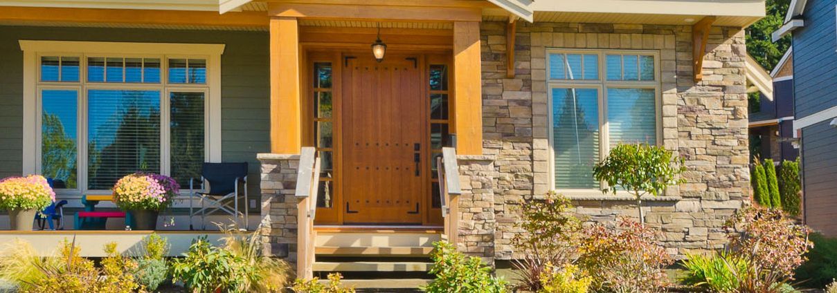 Front porch of a stone-and-wood house with blue windows, potted flowers, and steps leading to a wooden door.