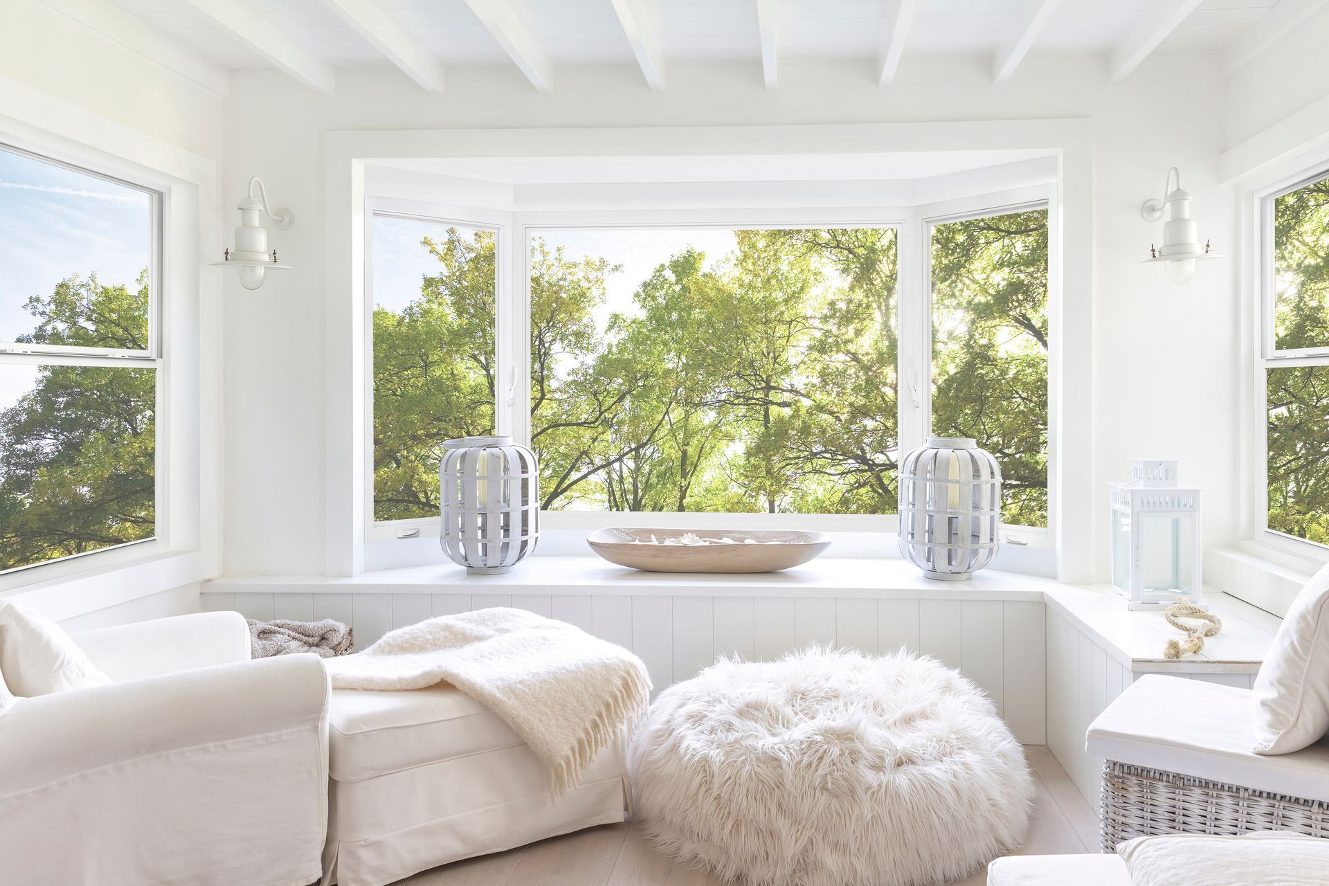 Bright white sunroom with cushioned seating and large windows overlooking green trees.