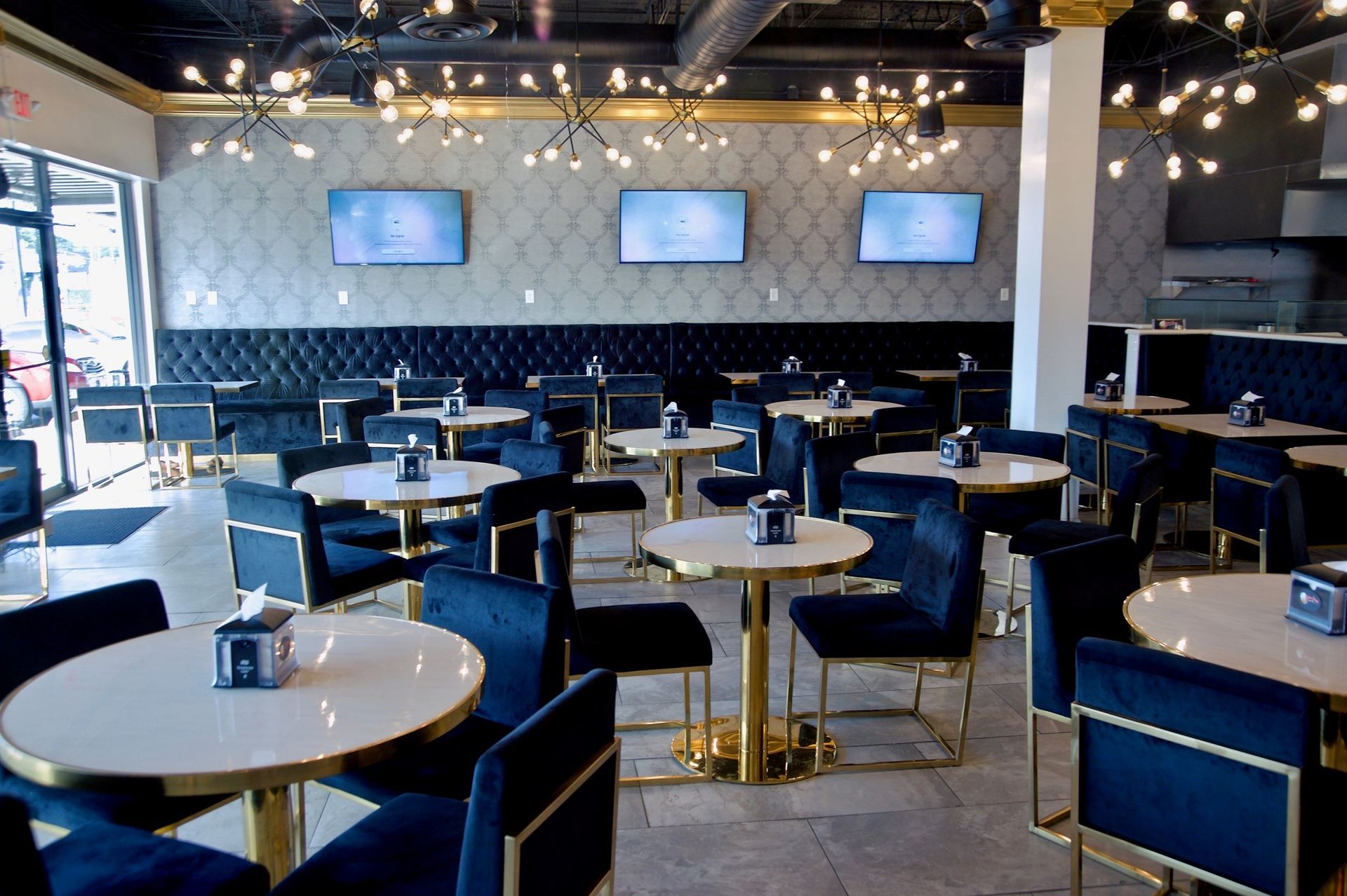 Restaurant interior with blue velvet chairs, gold tables and accents, and TVs on patterned wallpaper.