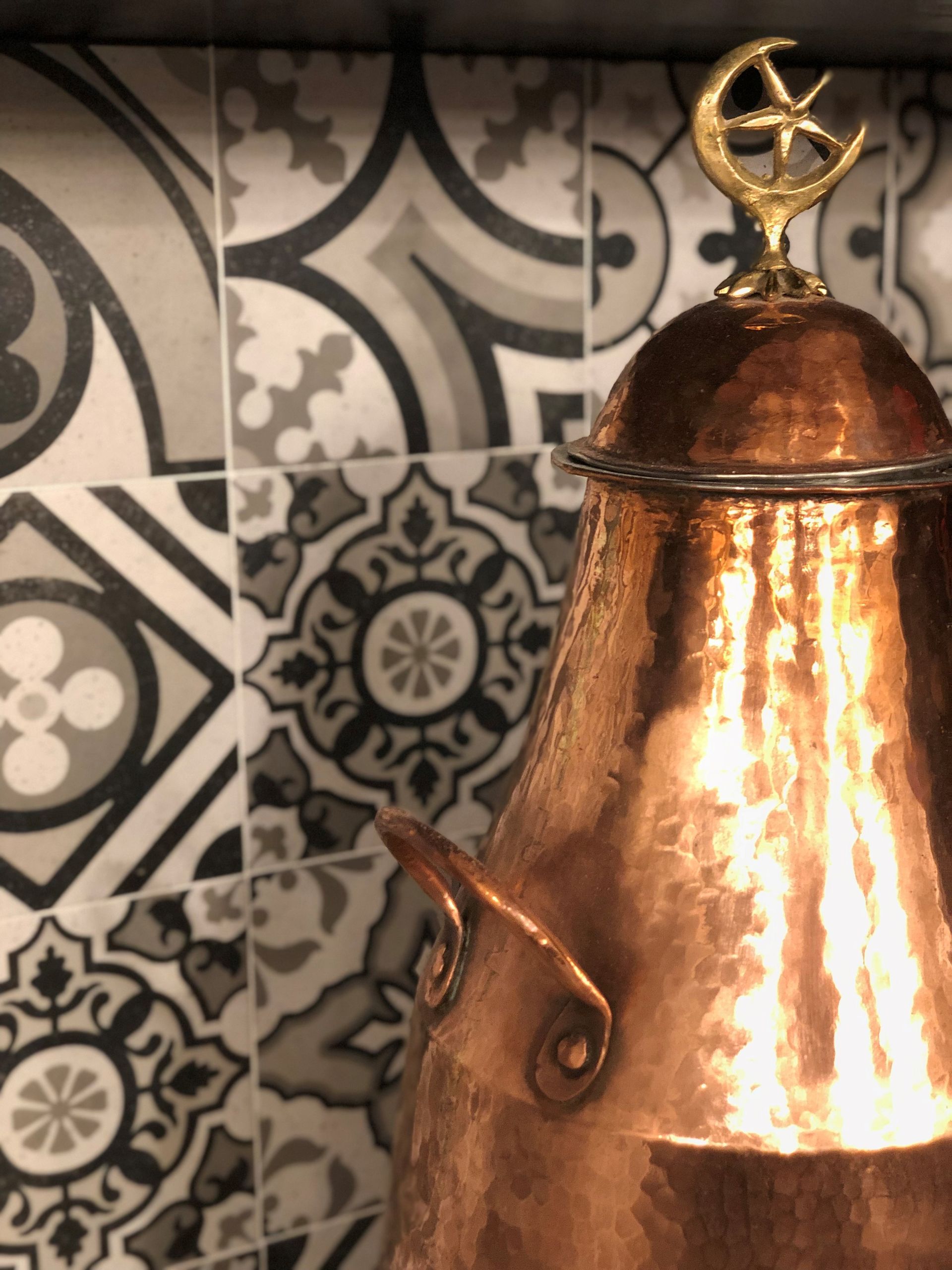 Copper teapot with ornate lid, against patterned tile background.