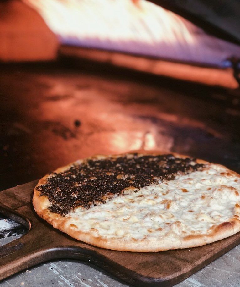Flatbread, half za'atar, half cheese, on a wooden paddle, near a hot oven.