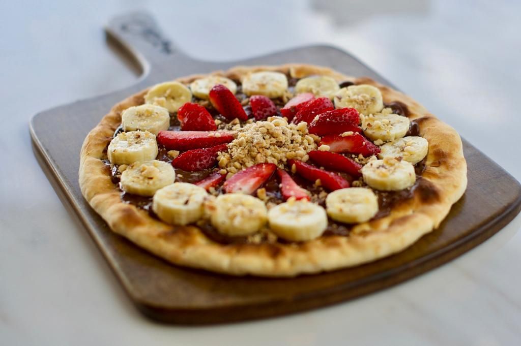 Nutella dessert pizza topped with sliced bananas, strawberries, and nuts on a wooden board.