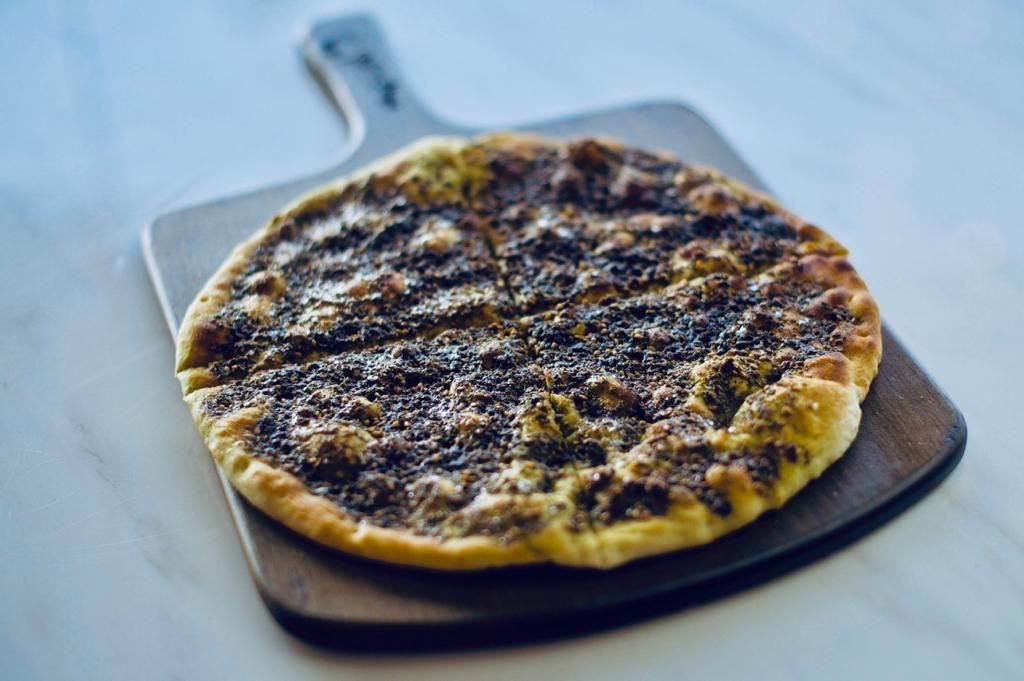 Round flatbread topped with dark za'atar, on a wooden serving board, set on a white surface.